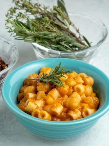 A teal bowl of pasta and chickpeas, with a sprig of rosemary on top, and a bowl of rosemary sprigs and red pepper flakes in the background.