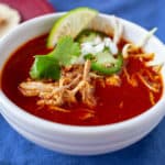 Bowl of Mexican Chicken Soup in Red Chile broth, with shreds of chicken, cilantro leaves, jalapeno slices, and diced onion on top
