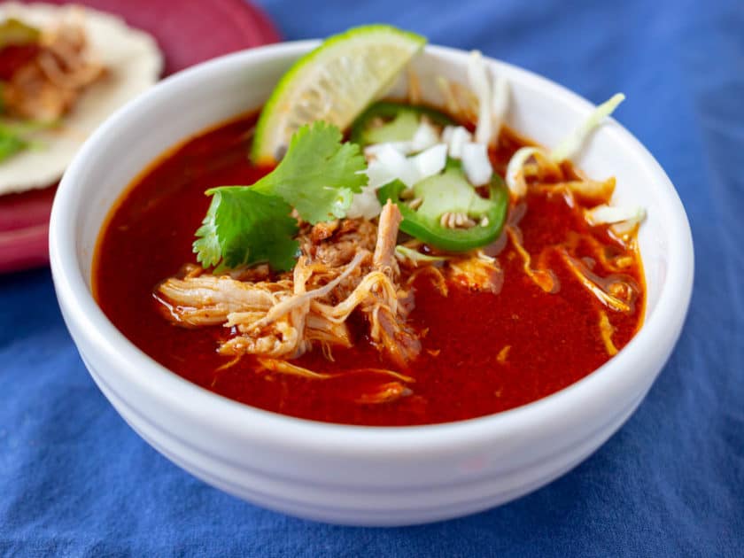 Bowl of Mexican Chicken Soup in Red Chile broth, with shreds of chicken, cilantro leaves, jalapeno slices, and diced onion on top
