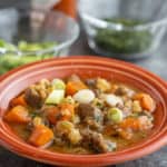 A bowl of beef brisket soup with carrots, green onions, and thyme, in an orange bowl on a slate-gray background