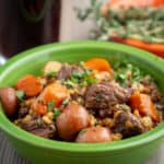 A green bowl full of beef stew with potatoes, carrots and barley; sprinkled with parsley and thyme leaves, with a glass of stout and a bowl of thyme in the background