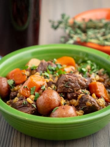 A green bowl full of beef stew with potatoes, carrots and barley; sprinkled with parsley and thyme leaves, with a glass of stout and a bowl of thyme in the background