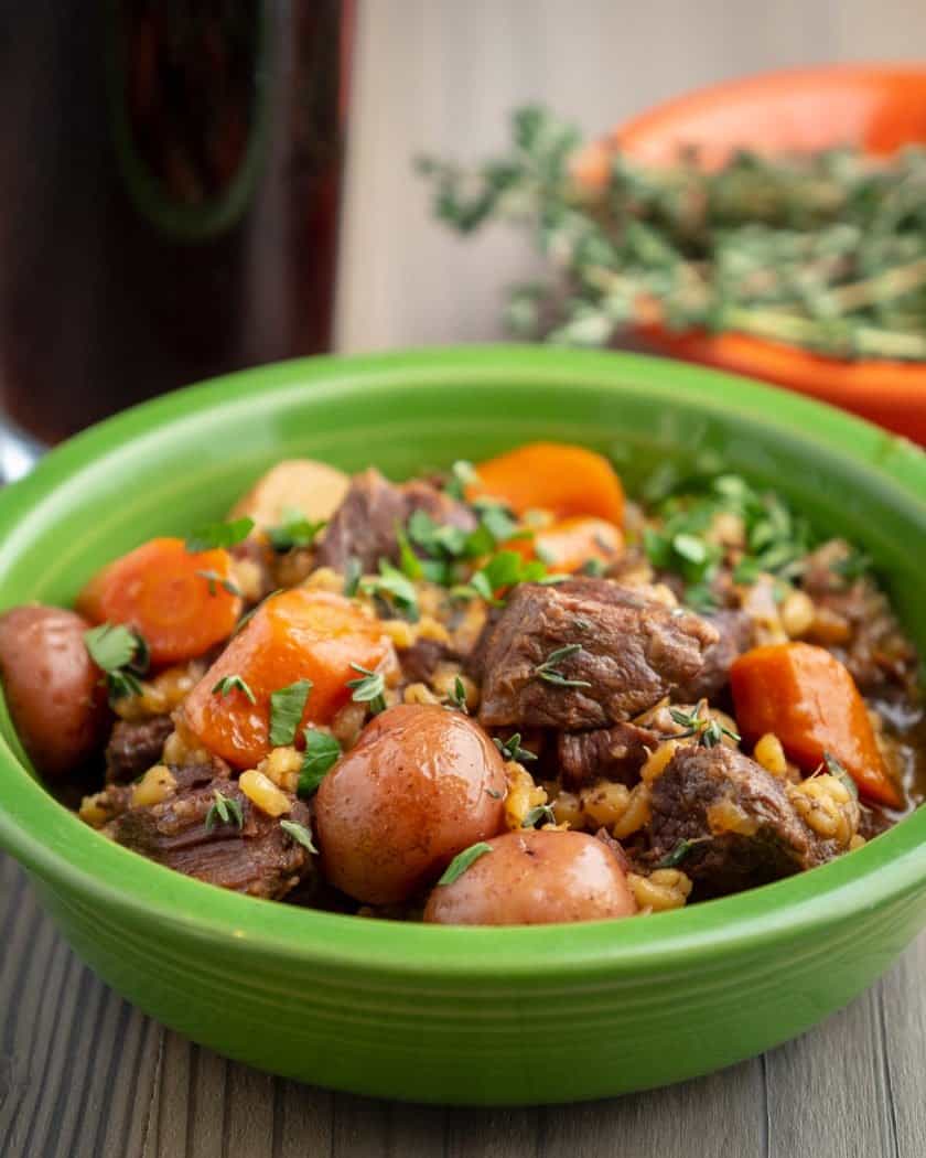 A green bowl full of beef stew with potatoes, carrots and barley; sprinkled with parsley and thyme leaves, with a glass of stout and a bowl of thyme in the background