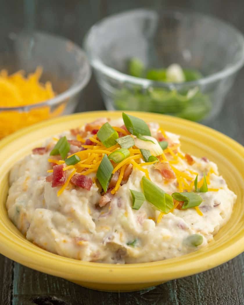 A yellow bowl of smashed red potatoes with bacon, shredded cheddar, and sliced green onions