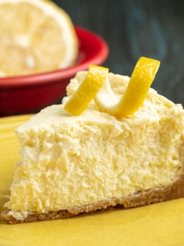A slice of lemon cheesecake with a lemon twist on top, on a yellow plate, with a lemon in a red bowl in the background