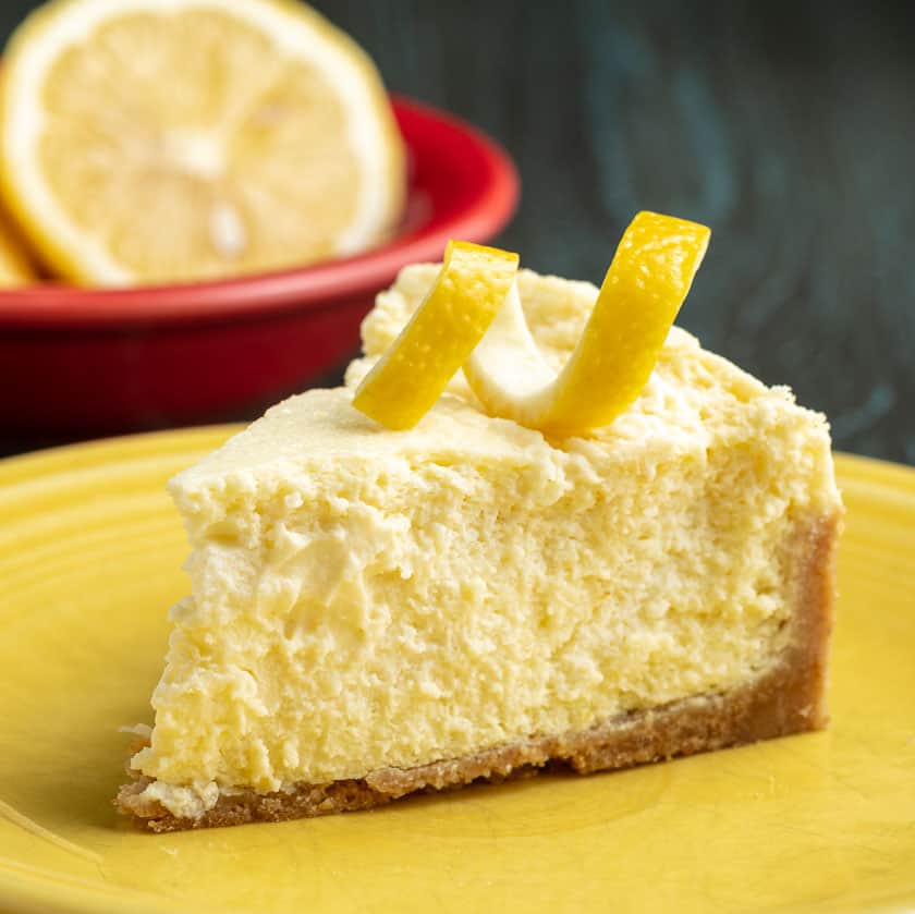 A slice of lemon cheesecake with a lemon twist on top, on a yellow plate, with a lemon in a red bowl in the background