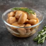 A bowl of cooked royal corona beans with a sprig of rosemary on top and on the table next to them.