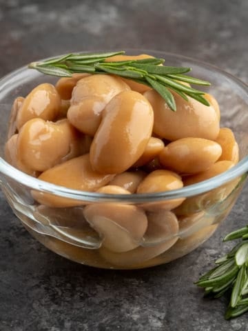 A bowl of cooked royal corona beans with a sprig of rosemary on top and on the table next to them.