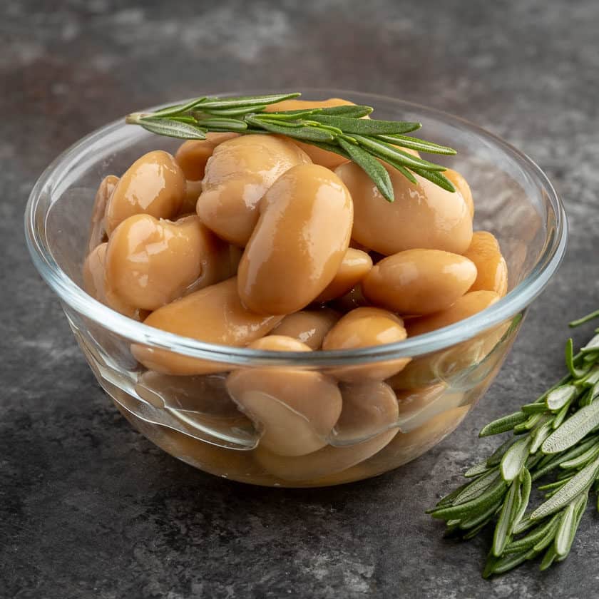 A bowl of cooked royal corona beans with a sprig of rosemary on top and on the table next to them.