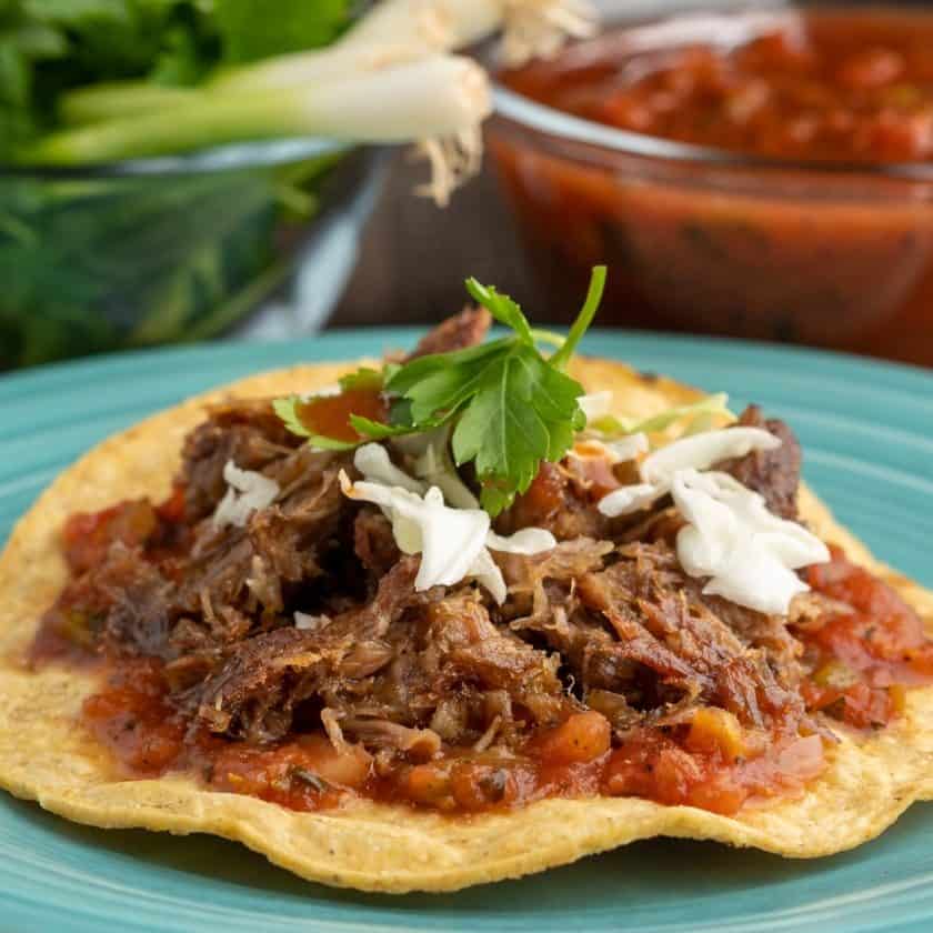 A tostada covered with salsa and crisped shredded pork, sprinkled with sliced cabbage and topped with a piece of cilantro, with green onions and salsa visible in the background