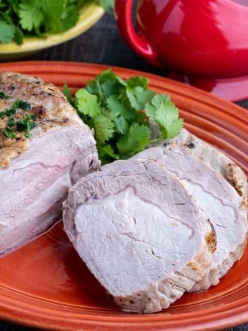 A sliced pork loin roast on an orange platter, with parsley and a gravy boat in the background