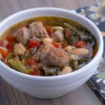 A white bowl of turkey sausage and kale soup, with beans and tomatoes, on a wooden table with a napkin.