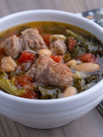 A white bowl of turkey sausage and kale soup, with beans and tomatoes, on a wooden table with a napkin.