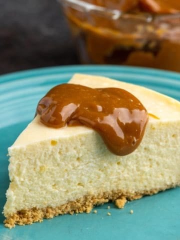 A piece of cheesecake with a dollop of dulce de leche on an aqua plate with a bowl of dulce de leche in the background