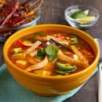 A yellow bowl of tortilla soup, with shreds of chicken, jalapeno peppers, tortilla strips, and cilantro, on a teal napkin, with lime wedges and chile peppers in the background.