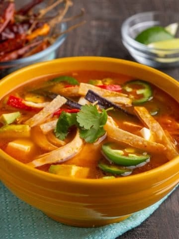 A yellow bowl of tortilla soup, with shreds of chicken, jalapeno peppers, tortilla strips, and cilantro, on a teal napkin, with lime wedges and chile peppers in the background.