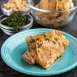 A chicken thigh, sprinkled with herbs, on a teal plate, in front of bowls of herbs and more chicken.