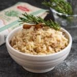 A white bowl of risotto with pork and cinnamon, topped by a rosemary sprig and cinnamon stick, with a bag of rice and more rosemary in the background