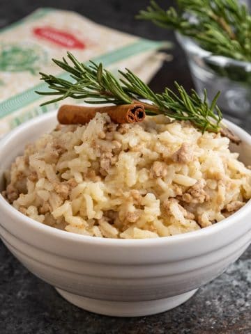 A white bowl of risotto with pork and cinnamon, topped by a rosemary sprig and cinnamon stick, with a bag of rice and more rosemary in the background