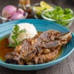 Short ribs with coconut curry sauce in a teal bowl with rice, and bowls of cilantro, shallots, limes, and curry paste in the background