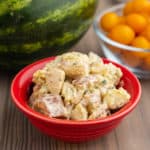 A red bowl of potato salad, on a wooden table, in front of a bowl of cherry tomatoes and a watermelon.