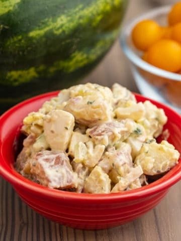 A red bowl of potato salad, on a wooden table, in front of a bowl of cherry tomatoes and a watermelon.