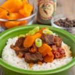 Beef stew with carrots, tomatoes, and green onions, on top of a bed of rice, in front of a bowl of scotch bonnet peppers, pickapeppa sauce, and allspice