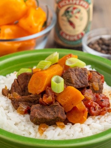 Beef stew with carrots, tomatoes, and green onions, on top of a bed of rice, in front of a bowl of scotch bonnet peppers, pickapeppa sauce, and allspice