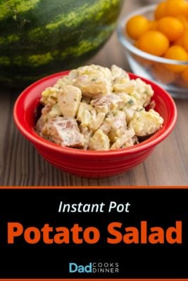A red bowl of potato salad, on a wooden table, in front of a bowl of cherry tomatoes and a watermelon.