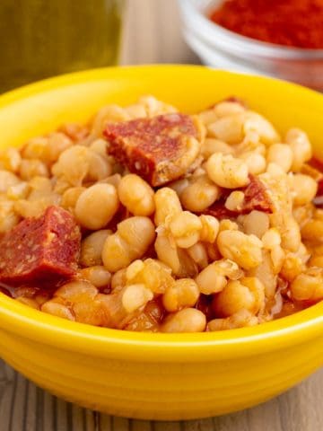 A yellow bowl of alubia blanca beans with chorizo and a red sauce, with olive oil and smoked Spanish paprika in the background.