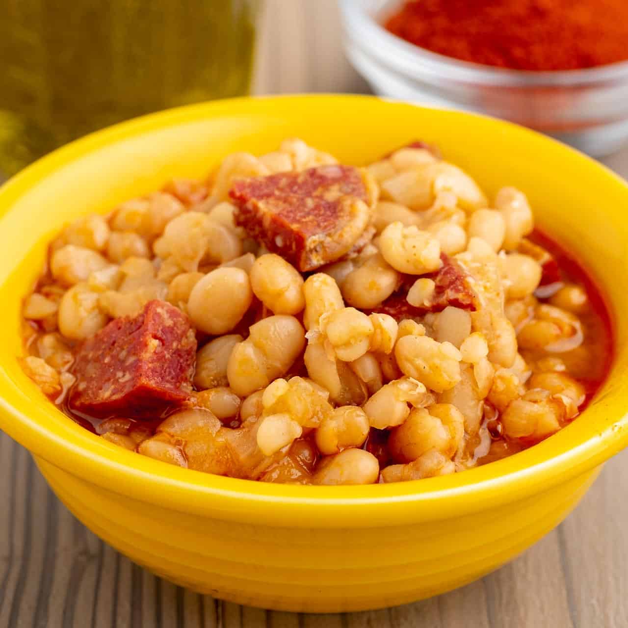 A yellow bowl of alubia blanca beans with chorizo and a red sauce, with olive oil and smoked Spanish paprika in the background.