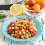 A plate of chickpeas with tomato lemon vinaigrette on a white table, with an Instant Pot, some minced basil, a half a lemon, and a bowl of tomatoes in the background.