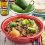 A red bowl of beef and tomatillo stew, with potatoes, sprinkled with sliced jalapeno, cilantro, and pepitas, with tortillas, jalapenos, and hot sauce in the background