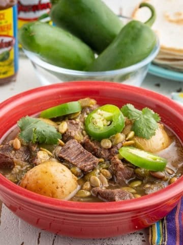 A red bowl of beef and tomatillo stew, with potatoes, sprinkled with sliced jalapeno, cilantro, and pepitas, with tortillas, jalapenos, and hot sauce in the background