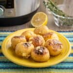 A plate of cooked potatoes, sprinkled with garlic and herbs, on a blue and yellow napkin with a pressure cooker, herbs, and a lemon in the background