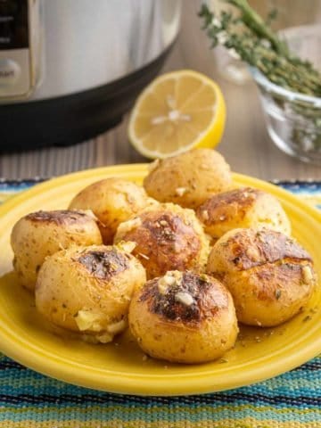 A plate of cooked potatoes, sprinkled with garlic and herbs, on a blue and yellow napkin with a pressure cooker, herbs, and a lemon in the background