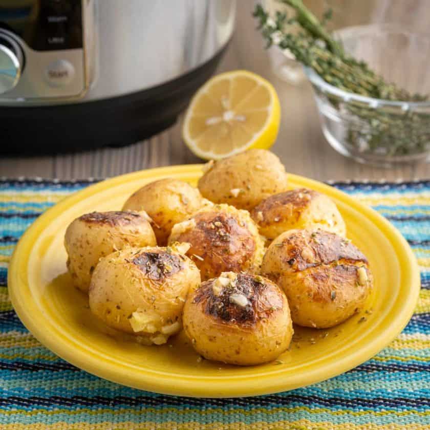 A plate of cooked potatoes, sprinkled with garlic and herbs, on a blue and yellow napkin with a pressure cooker, herbs, and a lemon in the background
