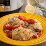 A plate of cooked chicken thighs, tomatoes, and onions, sprinkled with herbs, with a pressure cooker, herbs, and a lemon in the background