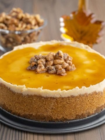 A maple cheesecake topped with maple syrup and candied walnuts. In the background is a maple leaf shaped jar of syrup and a bowl of walnuts.