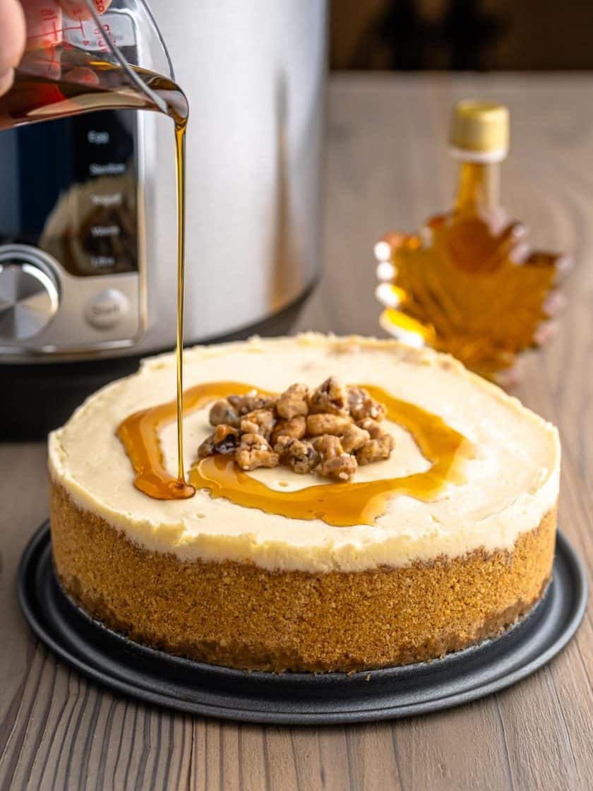 A cheesecake with maple syrup being drizzled on it, with an Instant Pot and a bottle of maple syrup in the background.