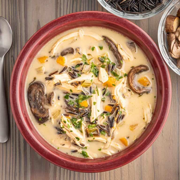 A creamy bowl of chicken and wild rice soup, with a spoon, some mushrooms, and some uncooked wild rice peeking in at the edges.