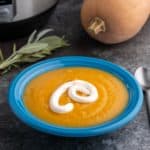 A blue bowl of squash soup with a drizzle of sour cream, with a bundle of herbs, a squash, and an Instant Pot in the background