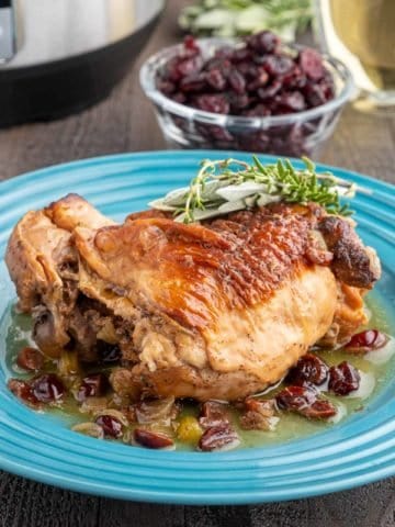 A turkey thigh on a bed of cranberries and onions and sauce, with a sprig of thyme and sage on top, and a cup of cranberries in the background