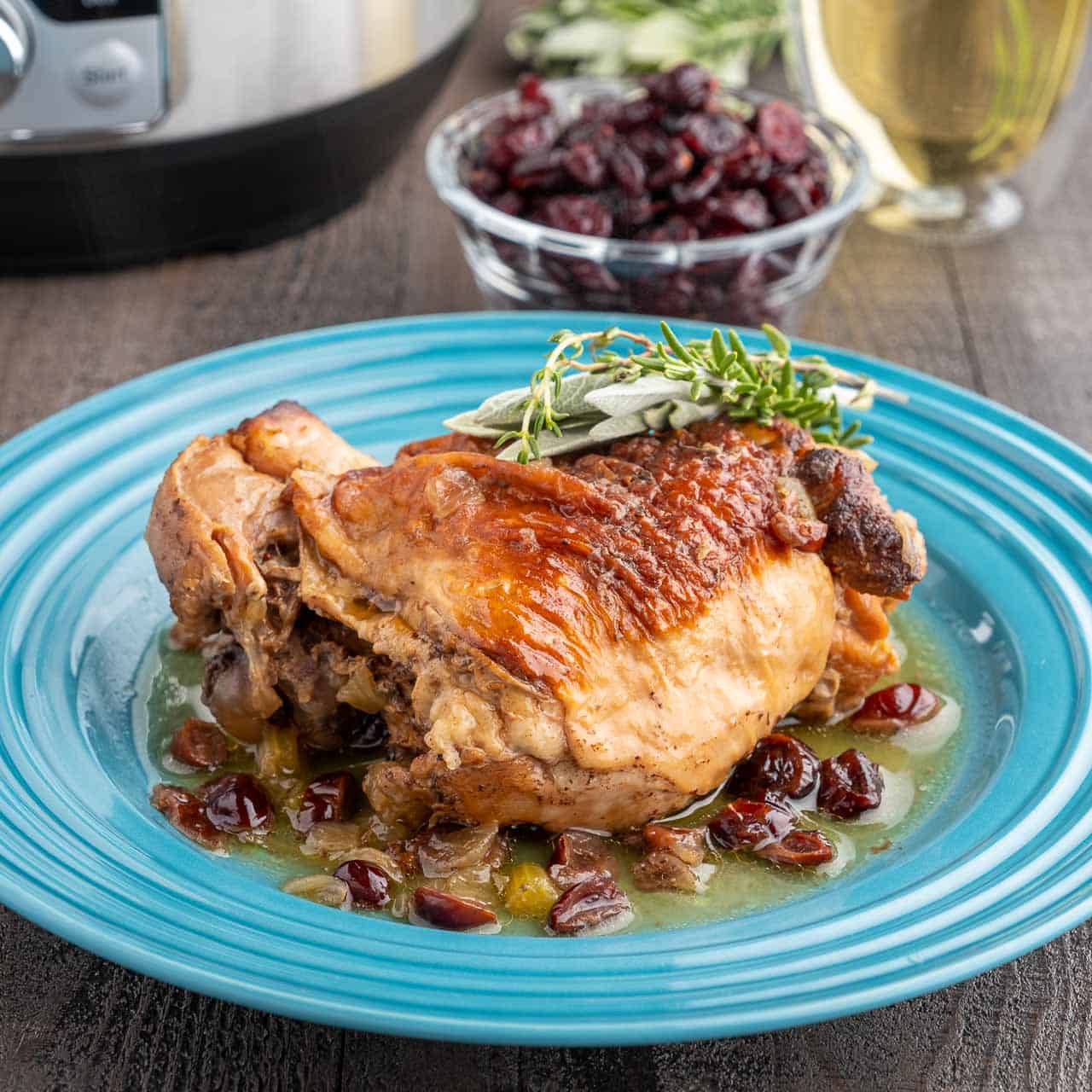 A turkey thigh on a bed of cranberries and onions and sauce, with a sprig of thyme and sage on top, and a cup of cranberries in the background