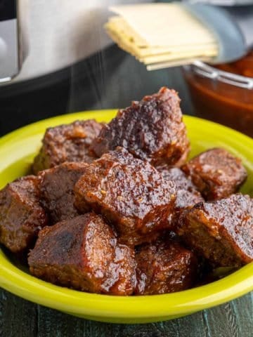 A bowl full of cooked brisket cubes, glazed with barbecue sauce, with an Instant Pot and a bowl of barbecue sauce in the background