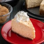 A slice of eggnog cheesecake on a red plate, with ginger snaps, an Instant Pot, and the rest of the cheesecake in the background