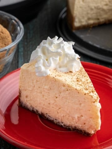 A slice of eggnog cheesecake on a red plate, with ginger snaps, an Instant Pot, and the rest of the cheesecake in the background