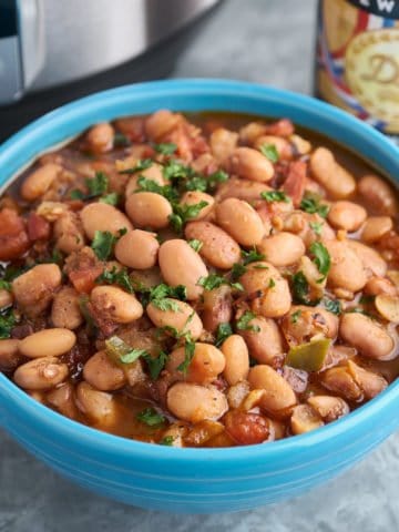 A blue bowl of borracho beans, pinto beans with bacon, tomato, and jalapeno, sprinkled with minced cilantro, with a bottle of beer and an Instant Pot in the background.