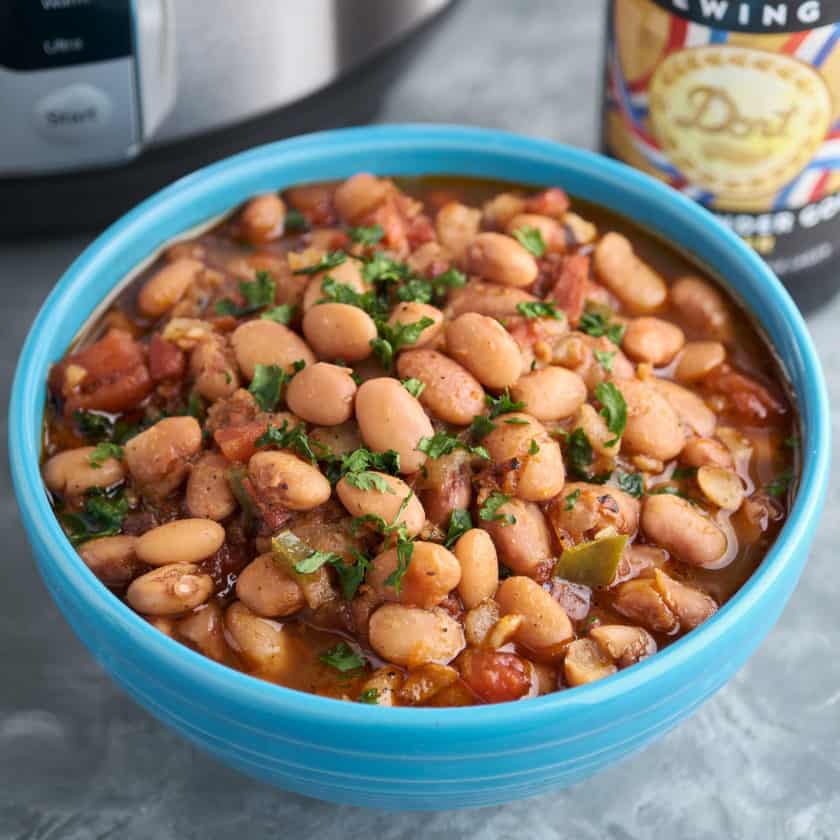 A blue bowl of borracho beans, pinto beans with bacon, tomato, and jalapeno, sprinkled with minced cilantro, with a bottle of beer and an Instant Pot in the background.