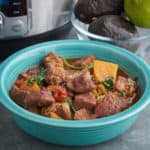 A bowl of pork stew with sweet potatoes, tomatoes, and black beans, with a sprinkle of cilantro, and an Instant Pot and bowl of avocados in the background